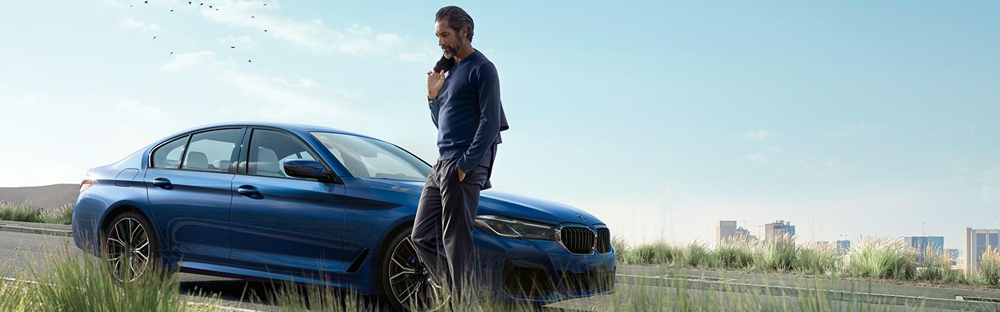 Man standing near a blue BMW 5 Series parked near grass.
